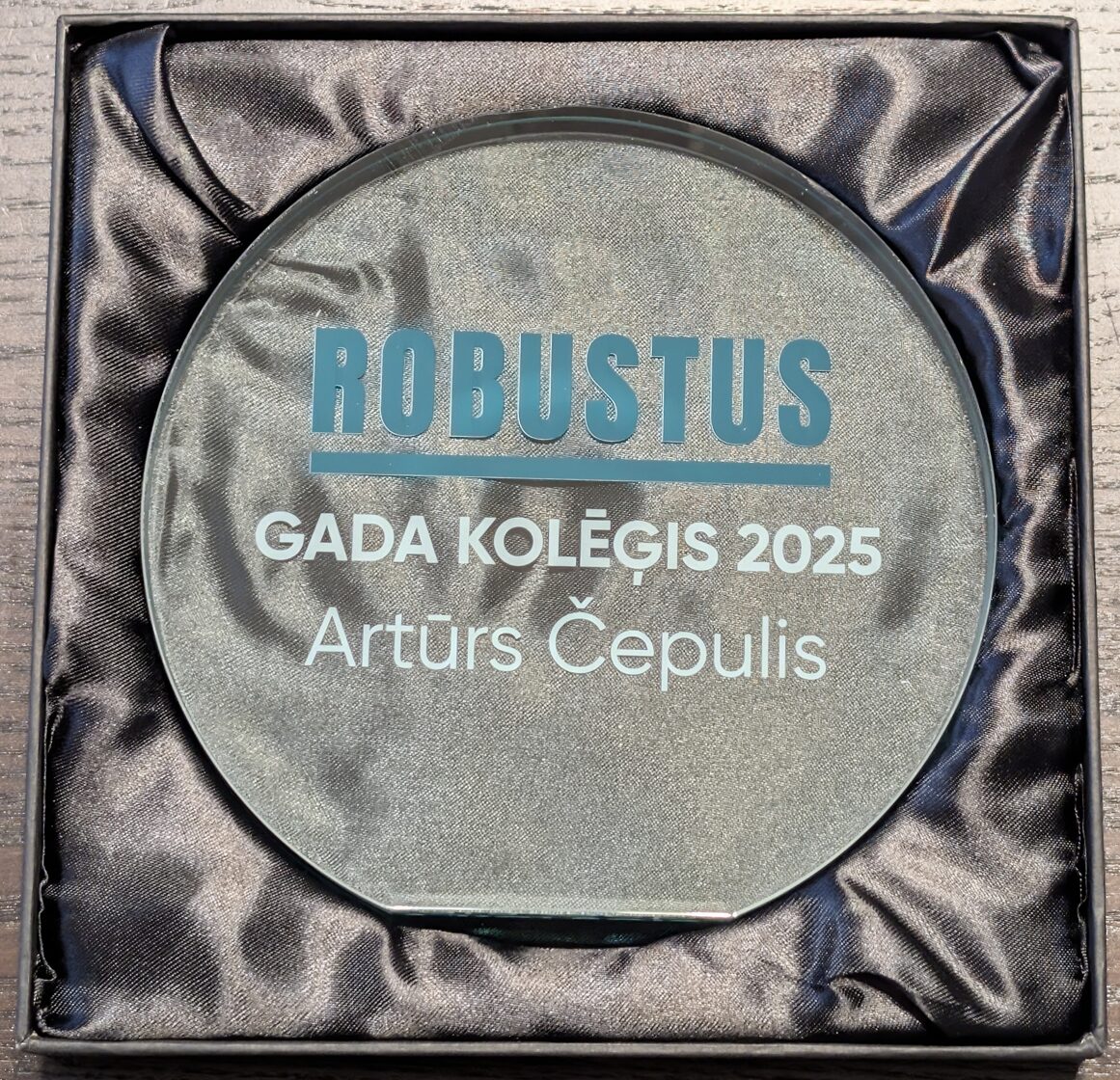 Apaļa stikla balva Gada kolēģis 2025 ar zilu Robustus logotipu kastītē.
